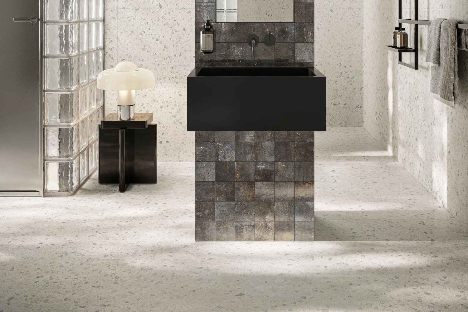 Sant Agostino Glow Light als helle Boden- und Wandfliese in feiner Terrazzo-Optik, kombiniert mit schwarzem Waschtisch und dunkler Mosaik-Akzentfläche im modernen Badezimmer