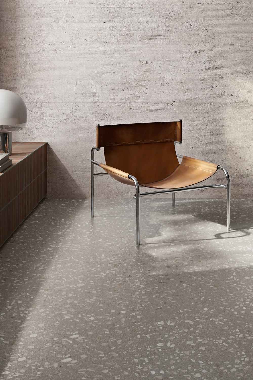 Sant Agostino Glow Grey als graue Bodenfliese in feiner Terrazzo-Optik mit hellen Steinfragmenten, kombiniert mit Lederstuhl und moderner Einrichtung