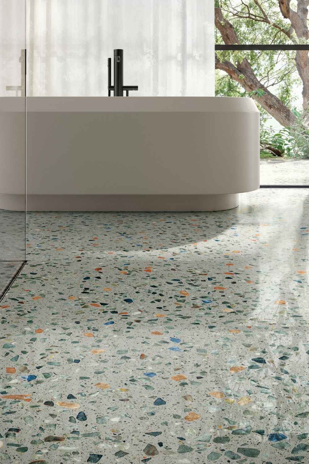 Sant Agostino Glow Plus Sage glänzend poliert als Bodenfliese in heller Terrazzo-Optik mit grünen, blauen, grauen und warmen Steinfragmenten vor einer freistehenden Badewanne