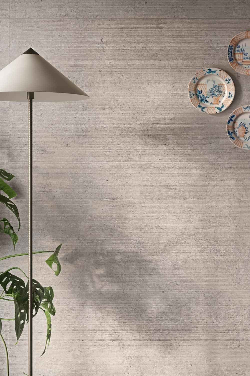 Sant Agostino Glow Concrete als großformatige Wandfliese in grauer Betonoptik mit feiner Struktur, kombiniert mit dekorativen Wandtellern, Stehleuchte und grüner Pflanze